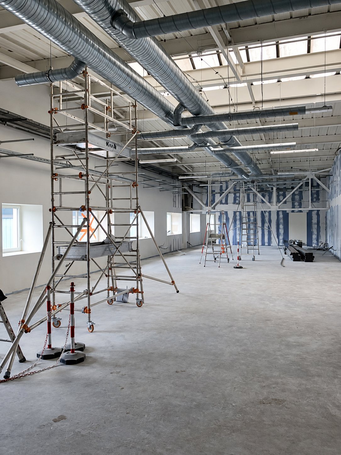 Vaste salle en travaux avec un échafaudage roulant en aluminium, des gaines de ventilation apparentes au plafond et des cloisons en cours d'installation.