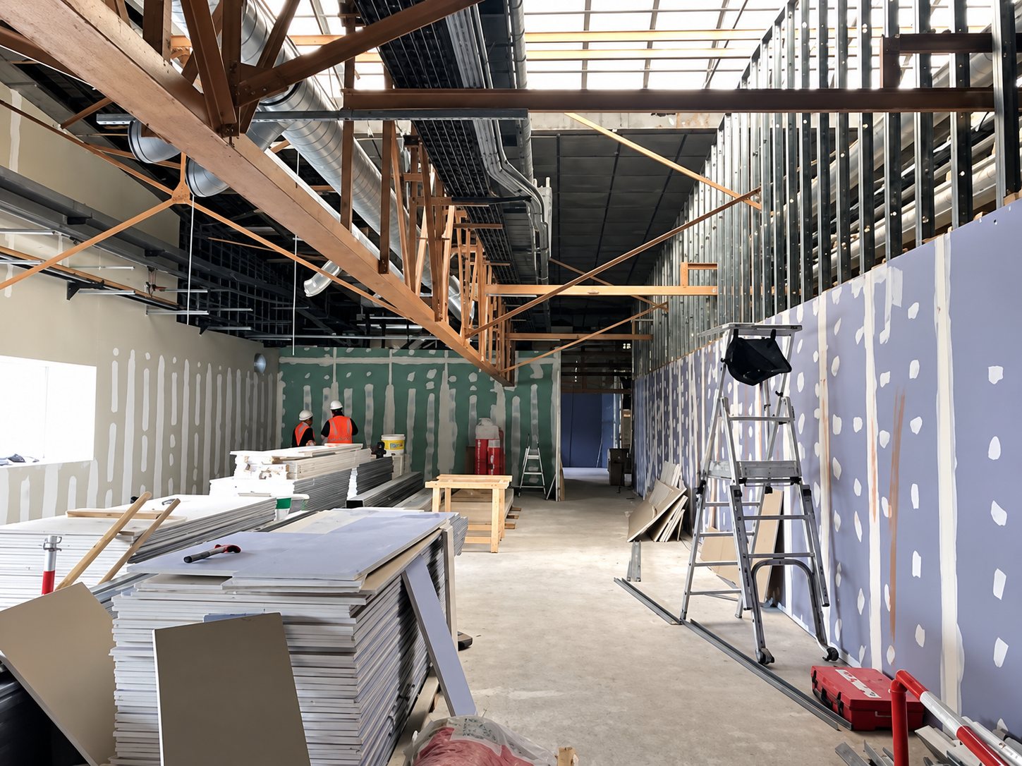 Vaste local en travaux avec piles de plaques de plâtre au premier plan, cloisons en cours de pose, une échelle et des ouvriers au fond.