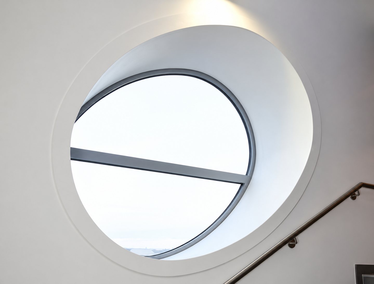 Une grande fenêtre circulaire encadrée de blanc laisse entrer la lumière dans une cage d'escalier avec une rampe métallique visible en bas à droite.