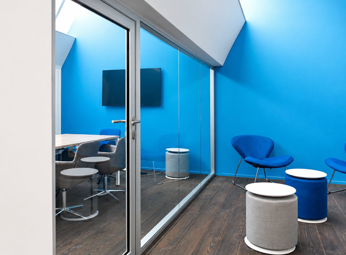 Une salle de réunion moderne aux murs bleus, séparée par une cloison vitrée, avec une table, des chaises grises, des fauteuils bleus et des poufs ronds sur un parquet sombre.